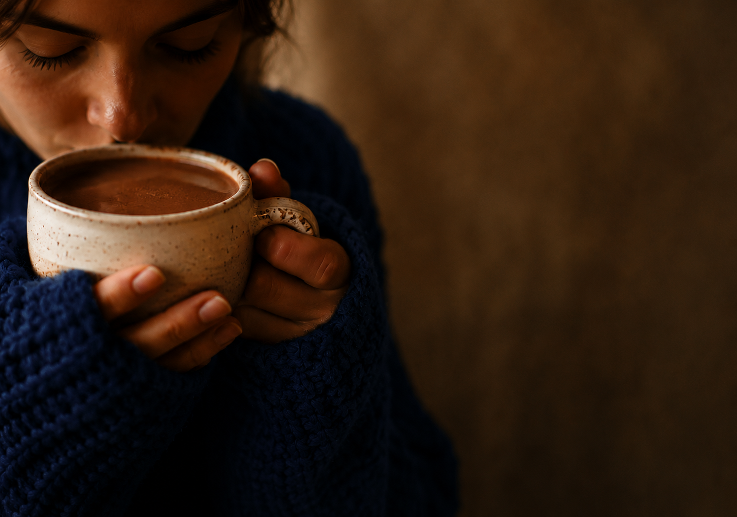 A close-up lifestyle shot of a person in a cozy dark blue knit sweater holding a warm ceramic mug filled with hot cocoa or coffee, with their face partially visible as they take a sip.